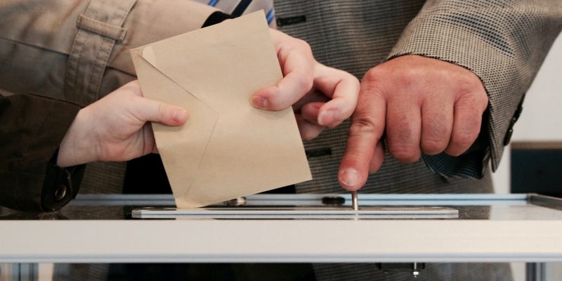 Ein Wahlumschlag wird in die Wahlurne eingeworfen, ein Wahlhelfer ist anwesend